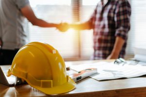 yellow safety helmet on workplace desk with construction worker team hands shaking greeting start up plan new project contract in office center at construction site, partnership and contractor concept